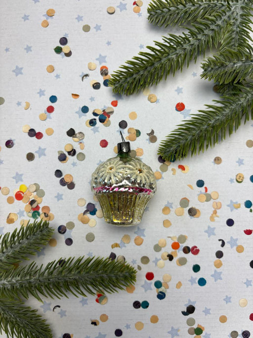 A decorative Christmas ornament with a cupcake-like design sits on a background of colorful confetti and pine branches.