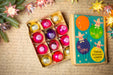 a box of colorful Christmas ornaments and a book with illustrations of Christmas ornaments on the cover, all placed on a table with a floral arrangement.