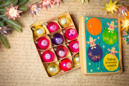 a box of colorful Christmas ornaments and a book with illustrations of Christmas ornaments on the cover, all placed on a table with a floral arrangement.