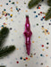 A pink ornament is placed on a white background, surrounded by colorful confetti and pine branches.