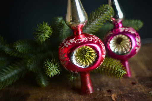 two vintage Christmas ornaments with red and pink colors, placed on a wooden surface next to some pine branches.
