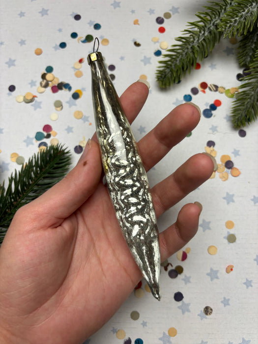 A hand holding a silver ornament with a textured surface, surrounded by colorful confetti and a pine branch.
