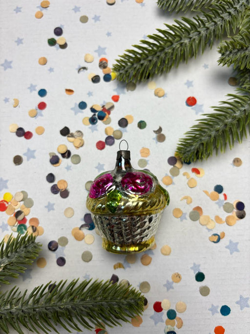 A glass ornament with a floral design sits on a white background, surrounded by colorful confetti and pine branches.