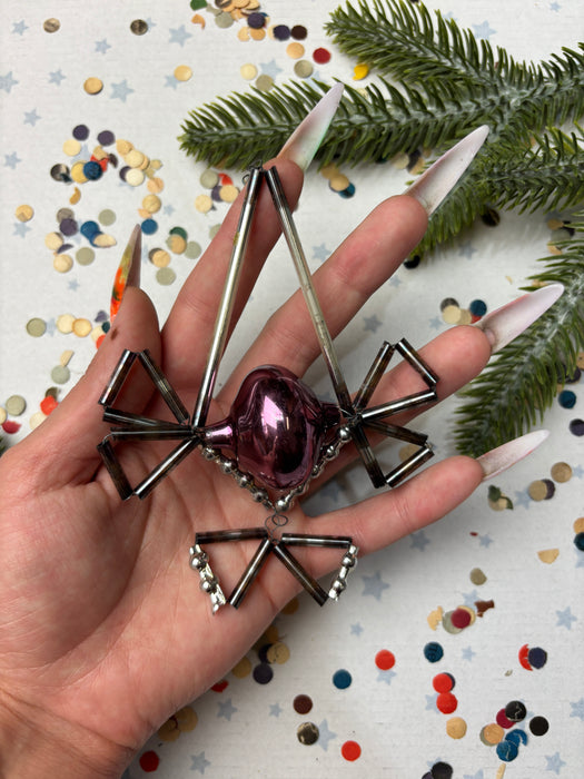 A hand holding a metallic ornament with a purple ball at the center, surrounded by various metallic shapes and a background of confetti.