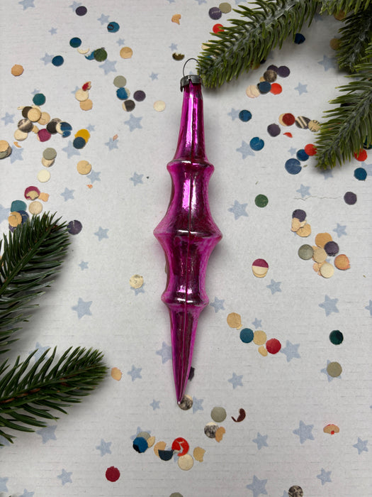 A pink ornament with a pointed end is surrounded by colorful confetti and pine branches on a white background.