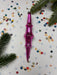 A pink ornament with a pointed end is surrounded by colorful confetti and pine branches on a white background.