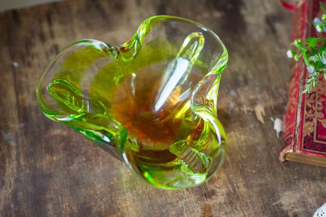 A green glass bowl with a curved shape sits on a wooden surface, with a red and gold book visible in the background.