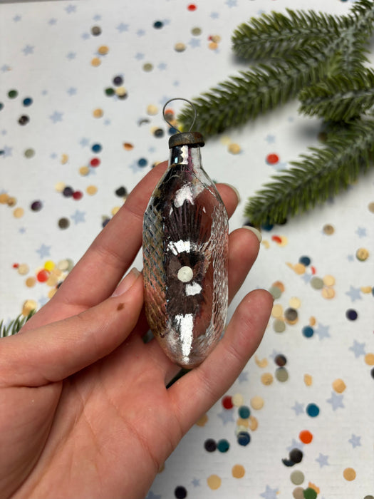 A hand holding a small, ornate glass ornament against a background of colorful confetti and a pine branch.