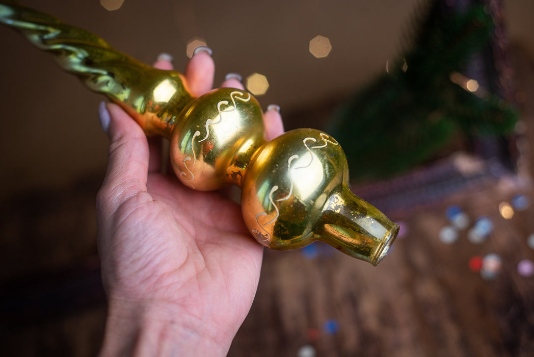 A hand holding a golden ornament with a floral design, against a blurred background with Christmas lights.