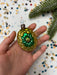A hand holding a small, ornate ornament with a green and gold design, surrounded by colorful confetti.