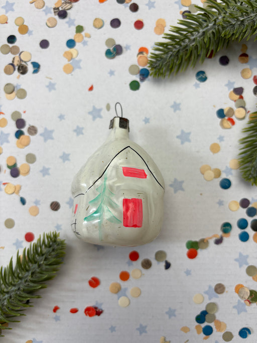A white ornament with red and green accents is placed on a white background with scattered confetti and green pine branches.