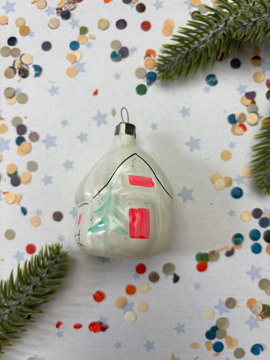 A white ornament with red and green accents is placed on a white background with scattered confetti and green pine branches.