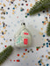 A white ornament with red and green accents is placed on a white background with scattered confetti and green pine branches.
