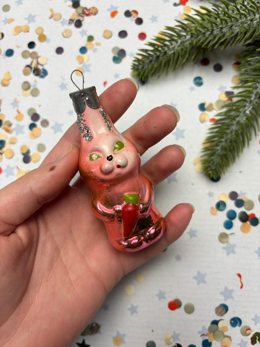 A hand holding a pink glass ornament with a carrot in it, surrounded by confetti and a pine branch.