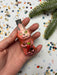 A hand holding a pink glass ornament with a carrot in it, surrounded by confetti and a pine branch.