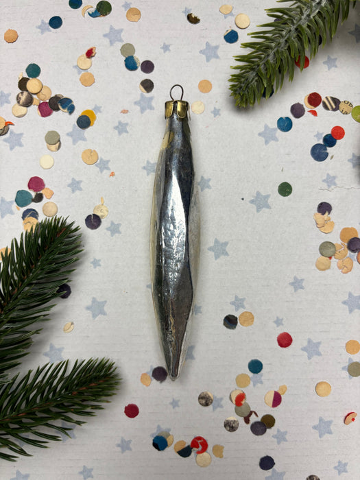 a silver ornament with a pointed end, surrounded by colorful confetti and a sprig of greenery.