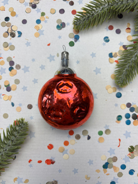 A shiny red Christmas ornament is placed on a white background, surrounded by colorful confetti and pine branches.