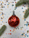 A shiny red Christmas ornament is placed on a white background, surrounded by colorful confetti and pine branches.