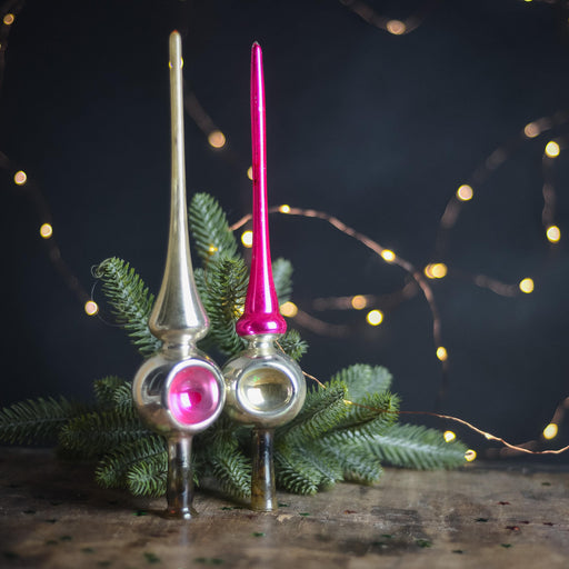 two ornaments, one silver and one pink, placed on a wooden surface with green pine branches and twinkling lights in the background.