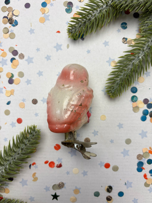 a pink and white bird ornament, surrounded by confetti and pine branches.