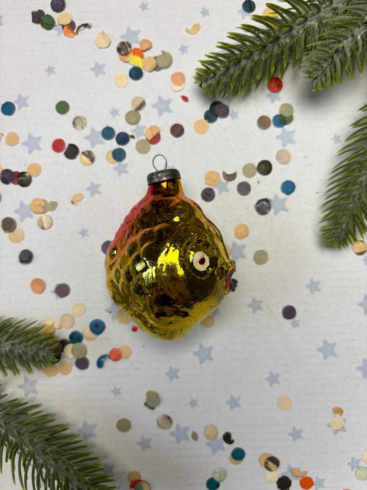 A shiny gold ornament resembling a fish is surrounded by colorful confetti and green pine branches on a white background.