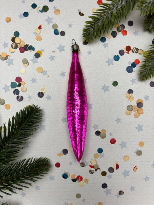 A pink ornament with a shiny, metallic appearance is displayed on a white background, surrounded by colorful confetti and pine branches.