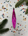 A pink ornament with a shiny, metallic appearance is displayed on a white background, surrounded by colorful confetti and pine branches.