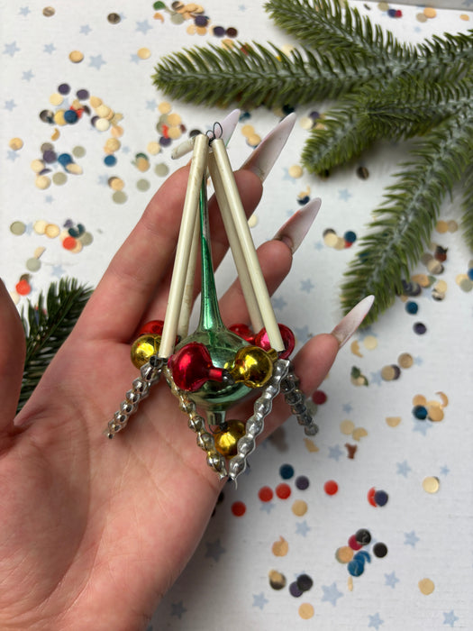 A hand holding a colorful ornament with bells and a green base, surrounded by confetti and a pine branch.