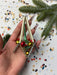 A hand holding a colorful ornament with bells and a green base, surrounded by confetti and a pine branch.