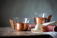 Two copper-colored mugs with wooden handles sit on a wooden surface, with a lace doily in the foreground.