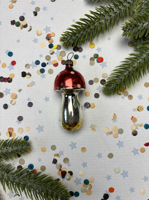 A red and silver ornament resembling a mushroom is placed on a white background with scattered confetti and pine branches.