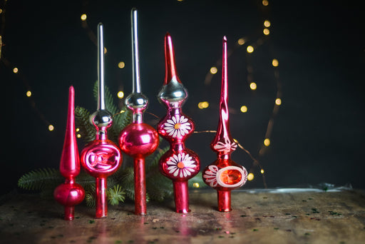A collection of colorful glass ornaments, including red and silver tops, are arranged on a wooden surface against a dark background with twinkling lights.