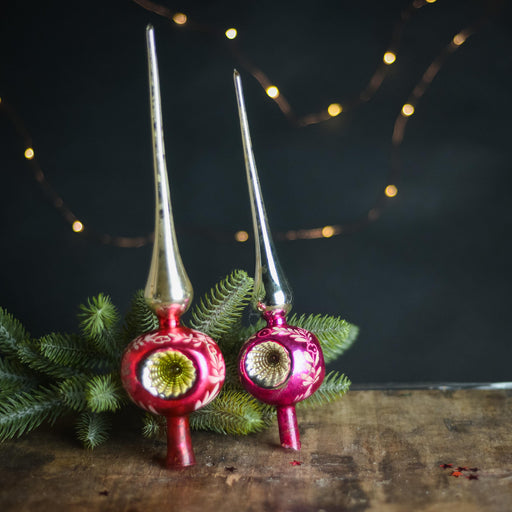 Two vintage Christmas ornaments with pointed tops and a green pine branch are displayed on a wooden surface, with twinkling lights in the background.