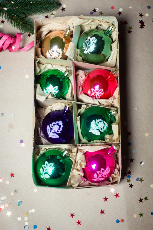 A box containing six colorful Christmas ornaments, including green, red, purple, and pink ornaments, surrounded by confetti.