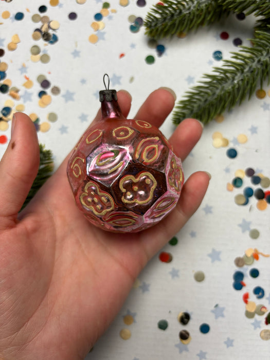 A hand holding a pink and gold ornament with a floral design, surrounded by confetti and a pine branch.