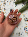 A hand holding a pink and gold ornament with a floral design, surrounded by confetti and a pine branch.