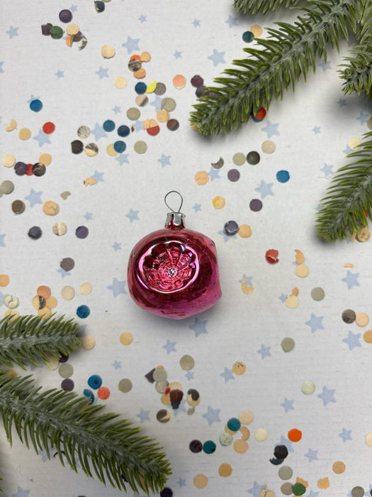A pink Christmas ornament with a red flower is surrounded by confetti and pine branches on a white background.