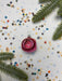 A pink Christmas ornament with a red flower is surrounded by confetti and pine branches on a white background.