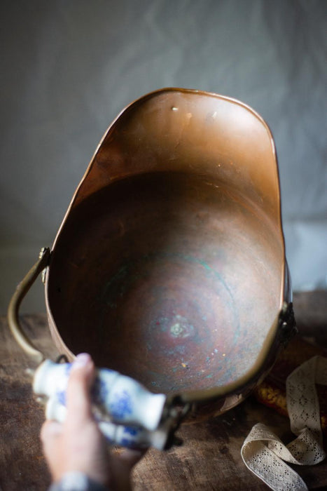A hand is holding a blue and white ceramic object, which appears to be a small bowl or dish, and is being used to clean a copper-colored metal object, possibly a pot or pan, on a wooden surface.