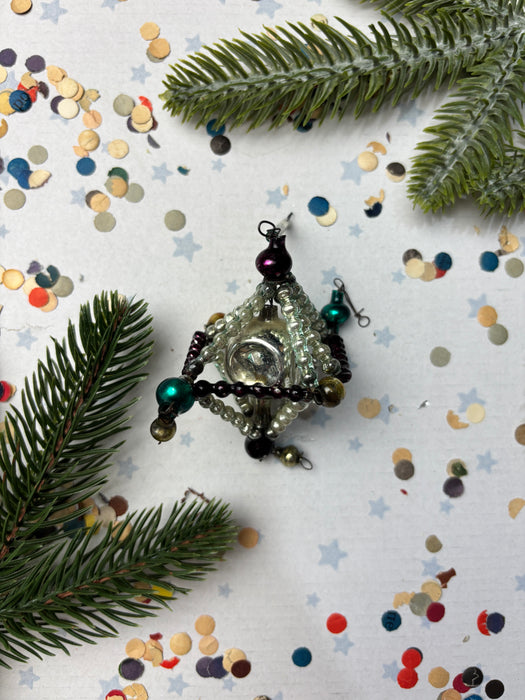 a Christmas ornament with a clear glass center, surrounded by a variety of colorful beads and a pine branch. The ornament is placed on a white background with scattered confetti.