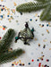 a Christmas ornament with a clear glass center, surrounded by a variety of colorful beads and a pine branch. The ornament is placed on a white background with scattered confetti.