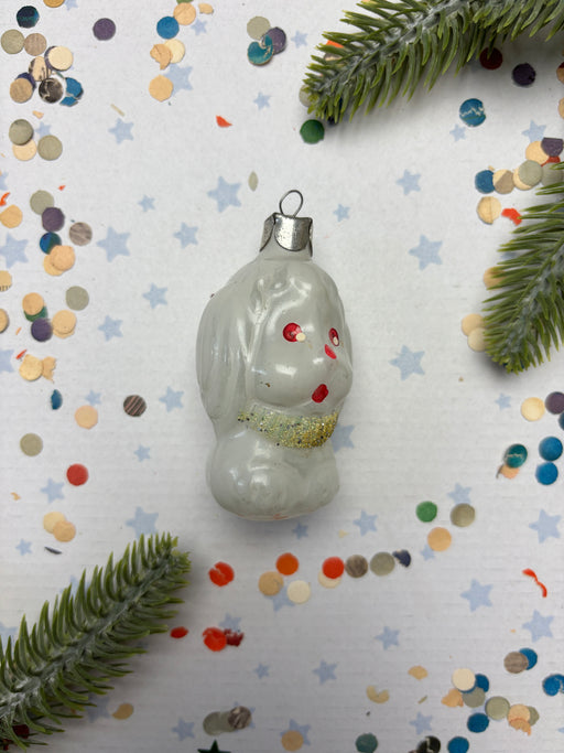 a white ornament with red and gold accents, surrounded by confetti and a sprig of greenery.