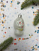 a white ornament with red and gold accents, surrounded by confetti and a sprig of greenery.