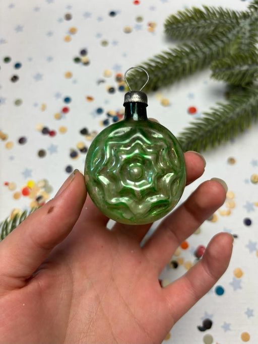 A hand holding a green glass ornament with a star-shaped design, against a background of confetti and a pine tree branch.