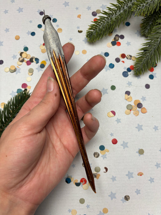 A hand holding a metallic object with a pointed end, surrounded by colorful confetti and a pine branch.