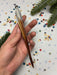 A hand holding a metallic object with a pointed end, surrounded by colorful confetti and a pine branch.