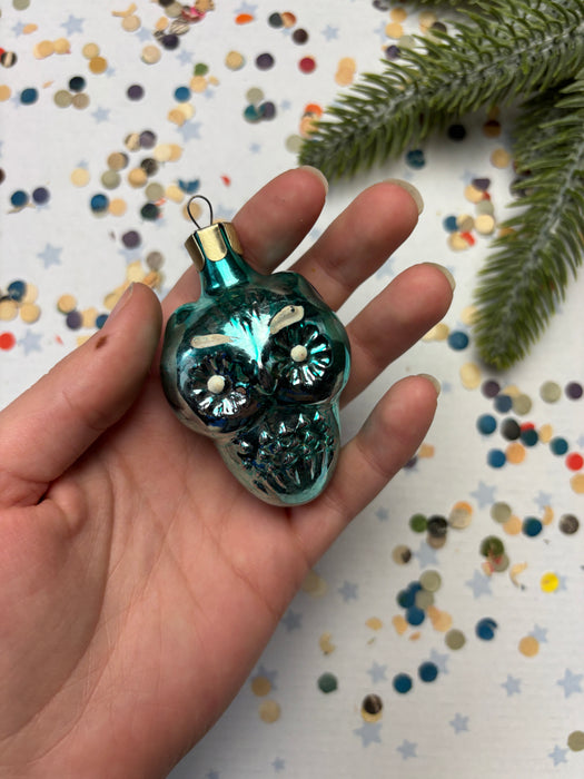 A hand holding a small, turquoise-colored ornament with a gold-colored stem, surrounded by colorful confetti on a white background.