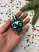 A hand holding a small, turquoise-colored ornament with a gold-colored stem, surrounded by colorful confetti on a white background.