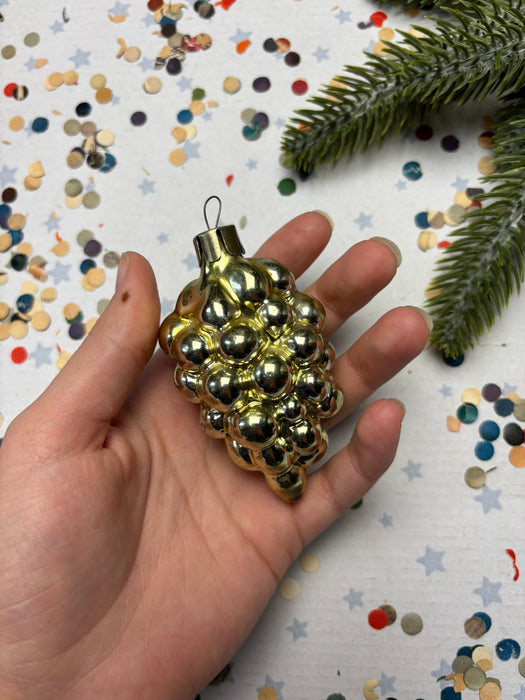 A hand holding a small, shiny gold ornament with a cluster of small, round balls. The hand is positioned against a background of confetti and a green pine branch.