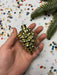 A hand holding a small, shiny gold ornament with a cluster of small, round balls. The hand is positioned against a background of confetti and a green pine branch.